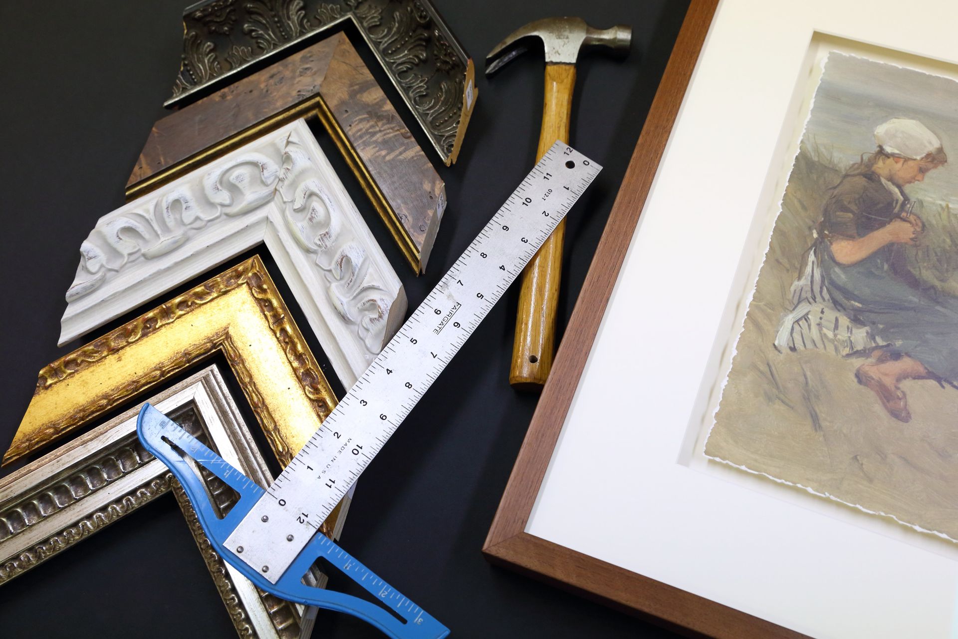 Frame moulding corner samples, a hammer, a ruler, and a partially matted print on a black work table