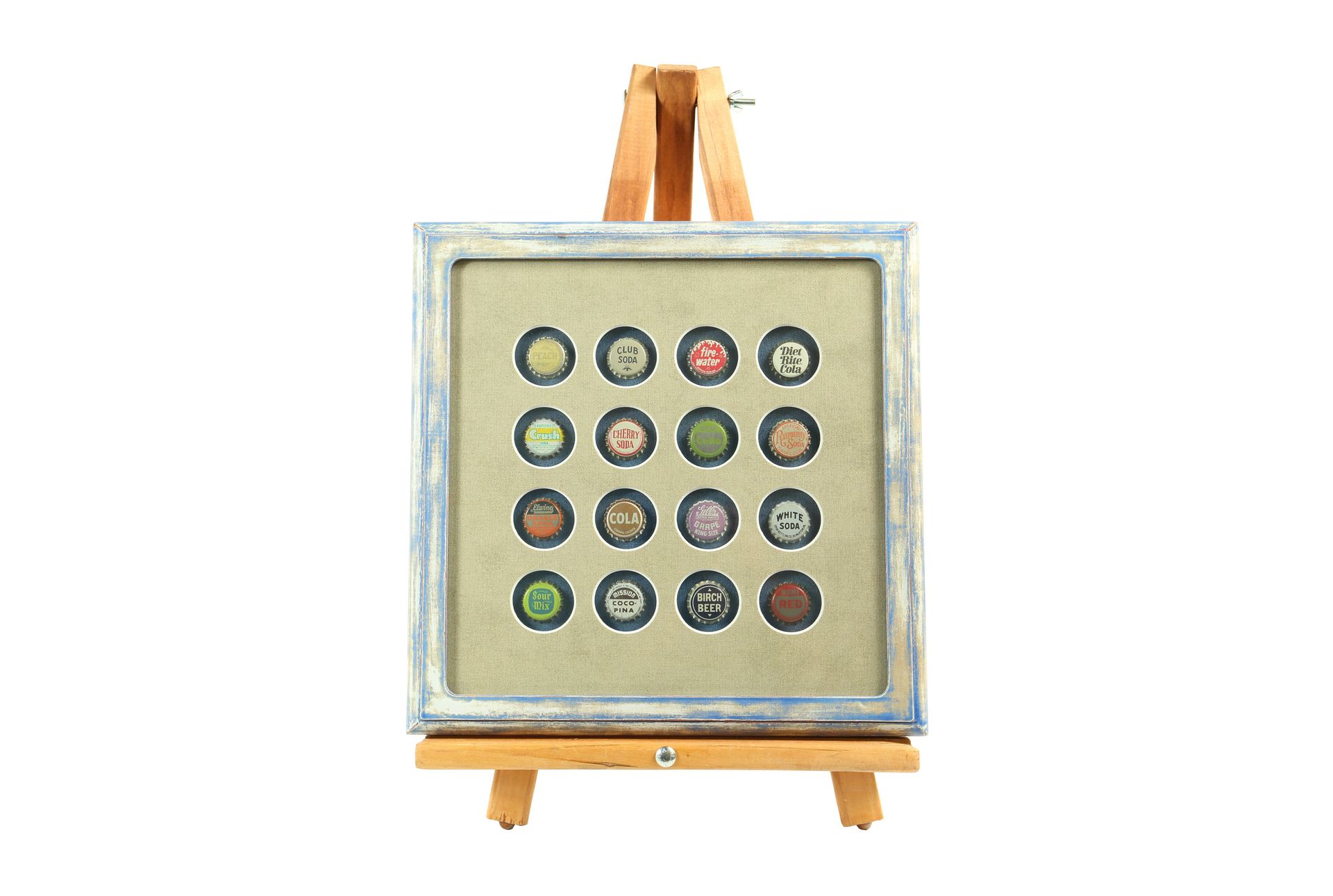 Sixteen vintage soda bottle caps arranged in a grid inside a distressed blue and gold shadow box on an easel